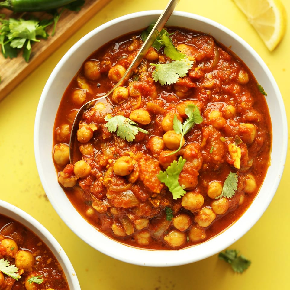 A white bowl of Indian chana masala, garnished with cilantro, with a spoon and lemon.