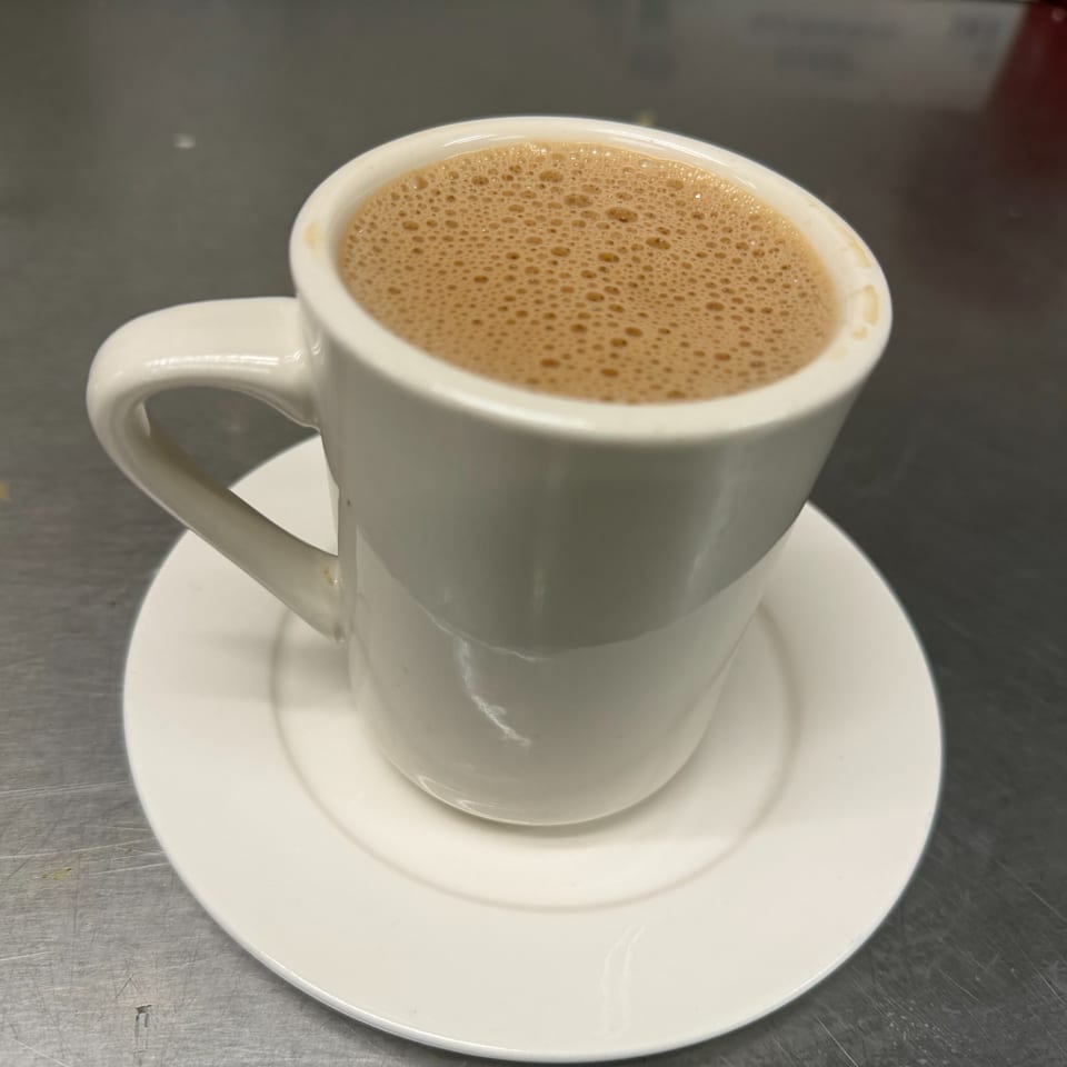 A white mug of frothy Indian chai tea sits on a white saucer on a metal surface.