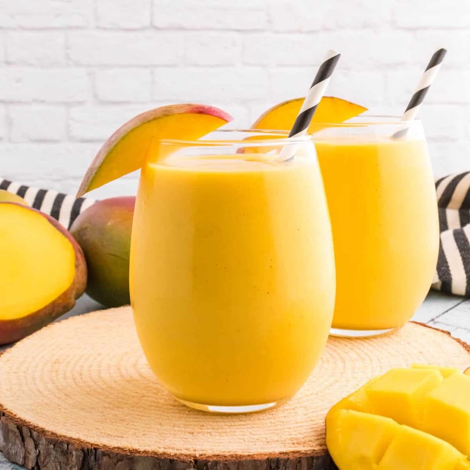 Two glasses of golden mango lassi with mango slices and striped straws on a wooden board.